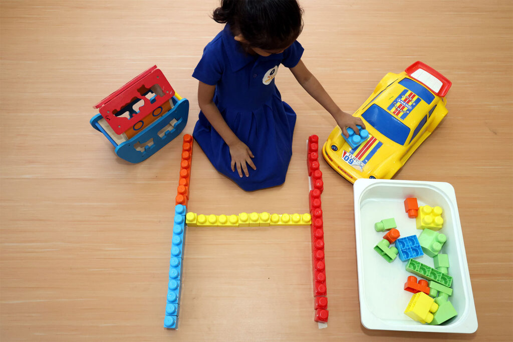 A young child in a blue uniform sits on a wooden classroom floor, playing with colorful building blocks and toy vehicles, demonstrating hands-on learning and creativity in a pre-primary school environment.