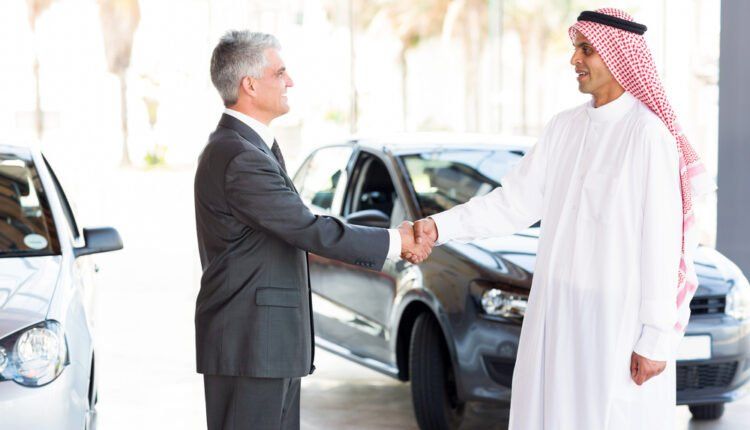 A car dealer in a traditional Arab outfit shaking hands with a customer beside a car at a dealership in Jordan.