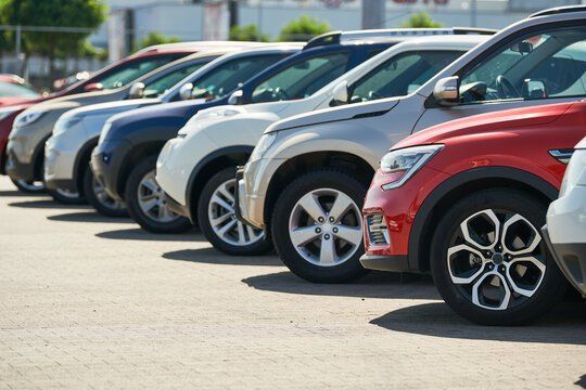 A row of parked used cars in a sunny lot, with a red SUV in front and white, silver and blue cars behind it.